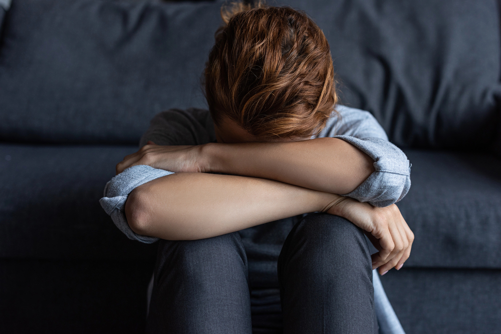 Person sitting on a dark sofa with head bowed and arms wrapped over their knees, conveying sadness or distress.