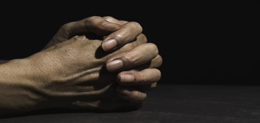 Two hands clasped together on a dark surface, fingers interlaced in a contemplative pose