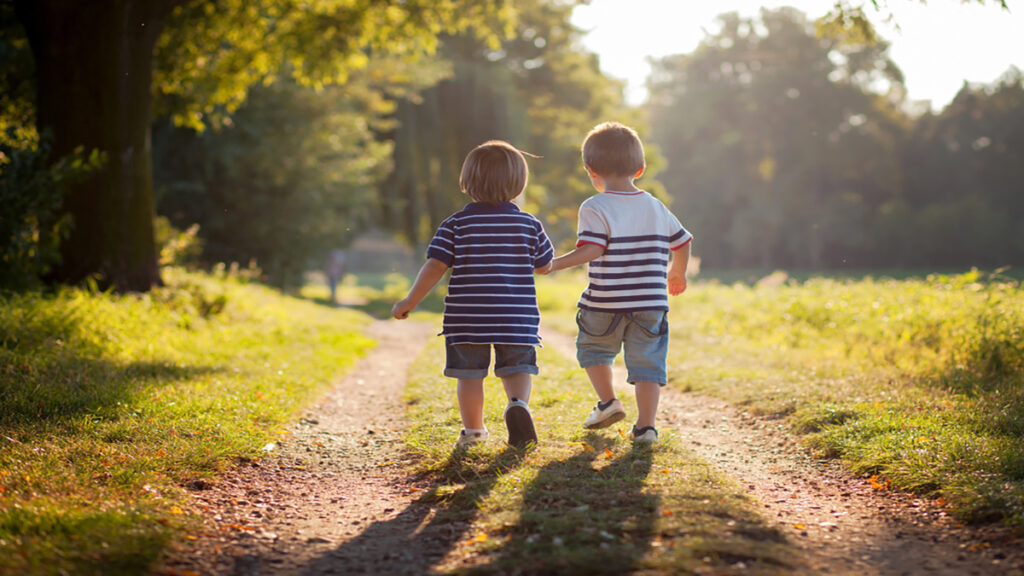 Two young children holding hands and walking down a sunlit park path.