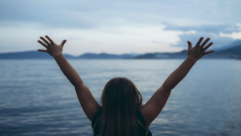Person facing away with arms raised, standing before a calm lake and distant mountains at sunset/dusk.