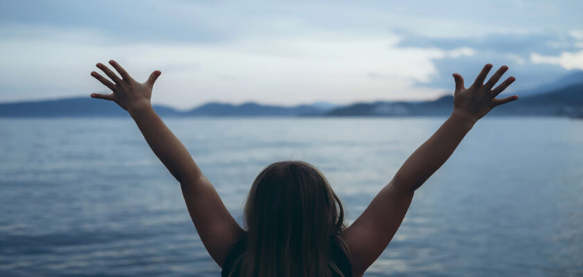Person facing away with arms raised, standing before a calm lake and distant mountains at sunset/dusk.