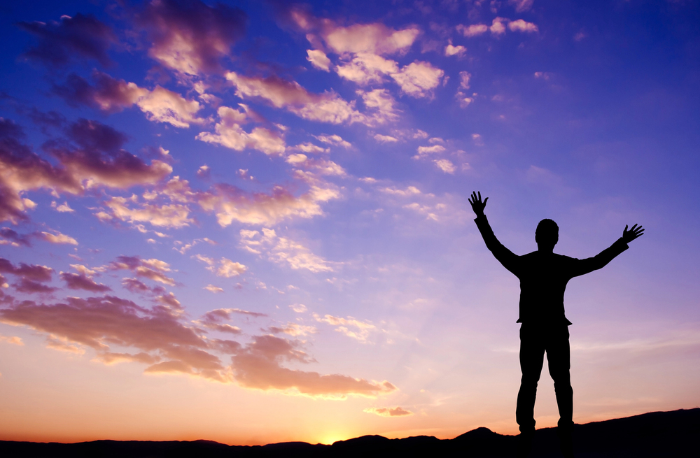 Silhouette of a person standing on a hill at sunset with arms raised toward the sky.