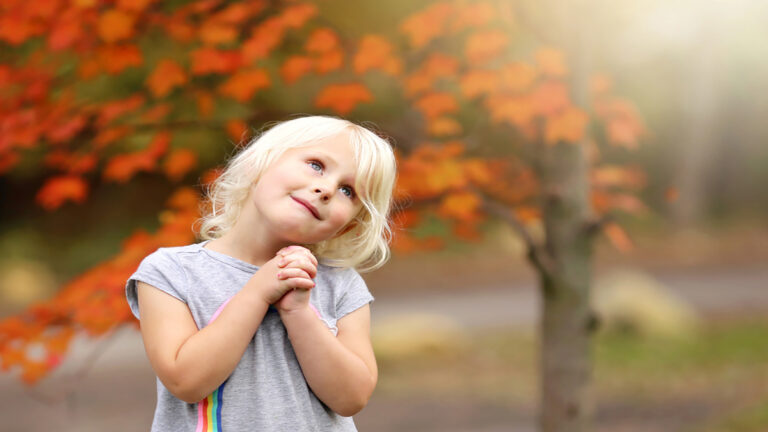 A smiling young girl with blonde hair in a gray shirt, clasping her hands and looking upward in a park with bright orange autumn leaves behind her.