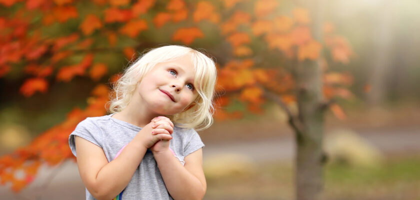 A smiling young girl with blonde hair in a gray shirt, clasping her hands and looking upward in a park with bright orange autumn leaves behind her.