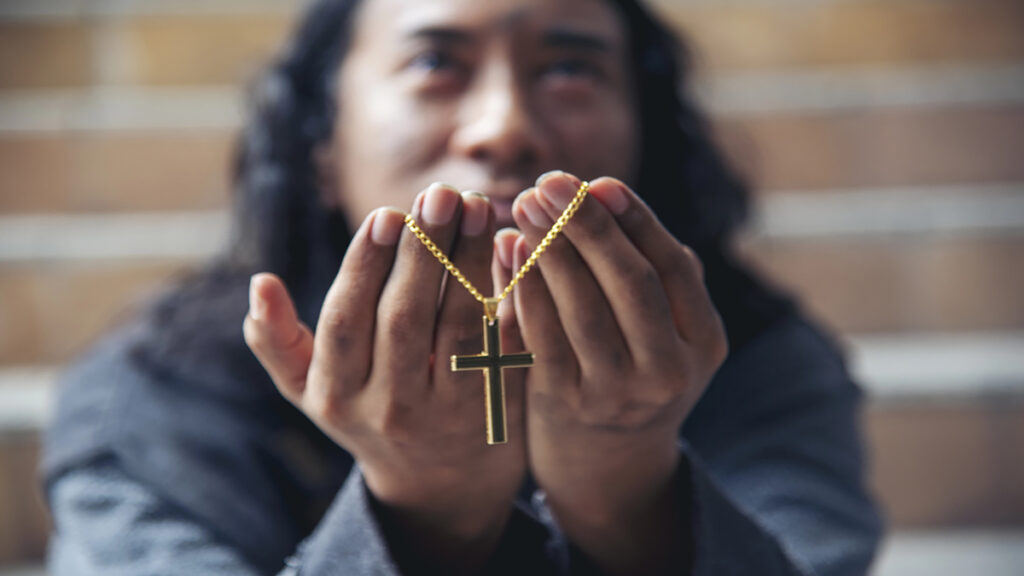 Person presenting a gold cross necklace held between cupped hands, with blurred brick steps in the background.