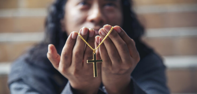Person presenting a gold cross necklace held between cupped hands, with blurred brick steps in the background.