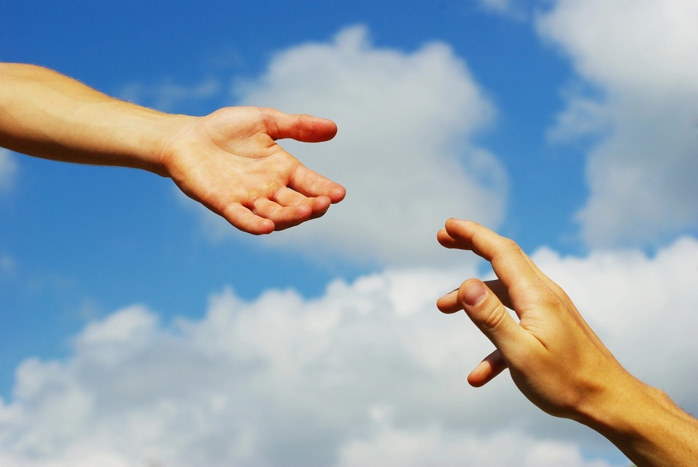 Two hands reaching toward each other against a blue sky with clouds, suggesting connection or help.