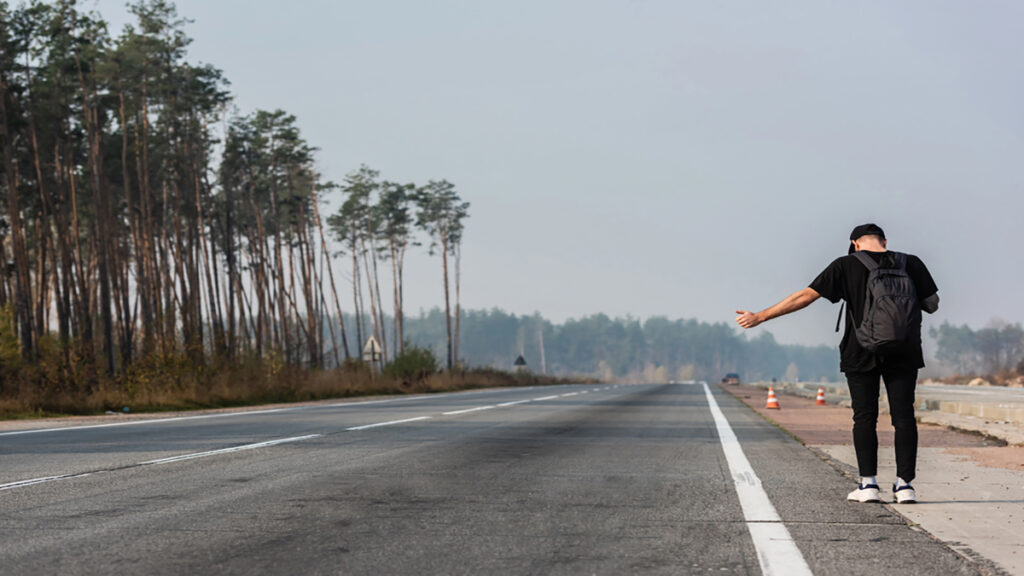 Man with a backpack stands on an empty road with arms outstretched, trees to the left and highway cones in the distance.