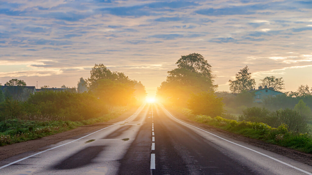 Two-lane rural road extends toward a sunrise, flanked by trees and bushes with golden light on the horizon.