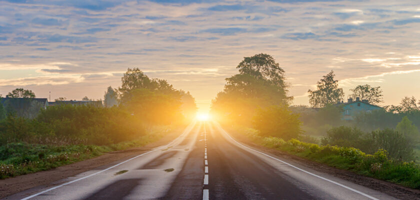 Two-lane rural road extends toward a sunrise, flanked by trees and bushes with golden light on the horizon.
