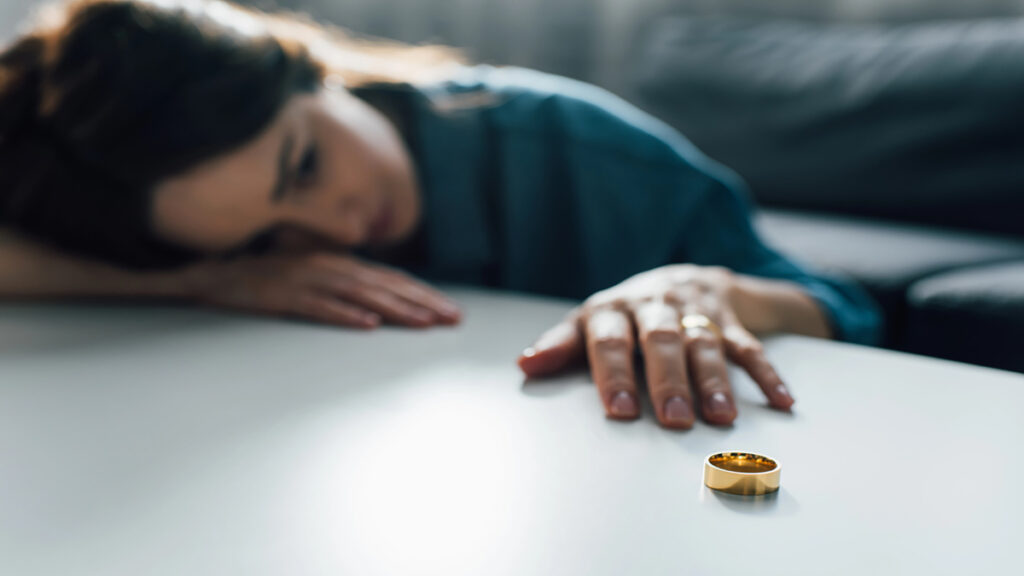 A person lies on a table nearby a gold wedding ring, suggesting loss or heartbreak.