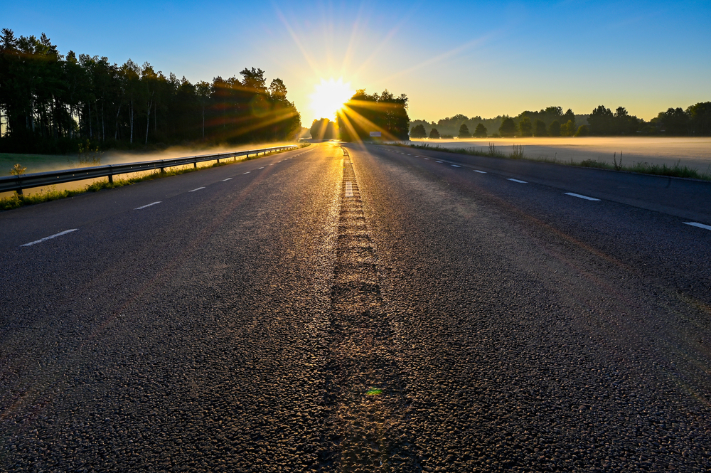 Two-lane road stretching toward the sunrise, with sunburst through trees and morning mist along both sides of the highway.