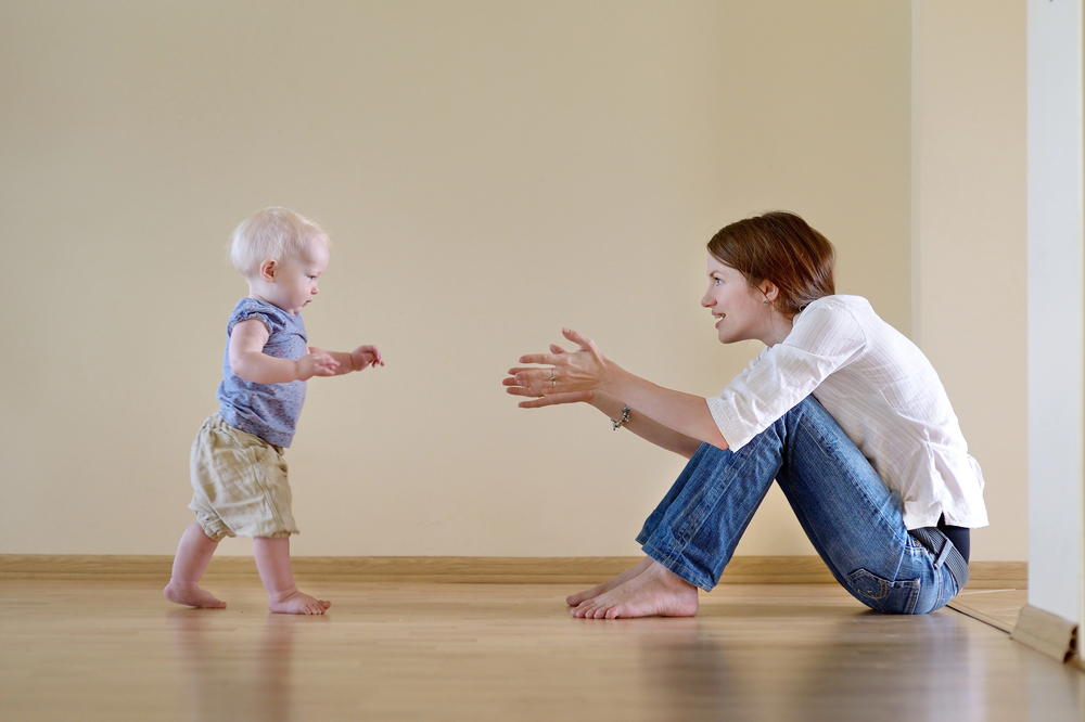Mother sitting on a light wooden floor, reaching toward her toddler who is standing and approaching.