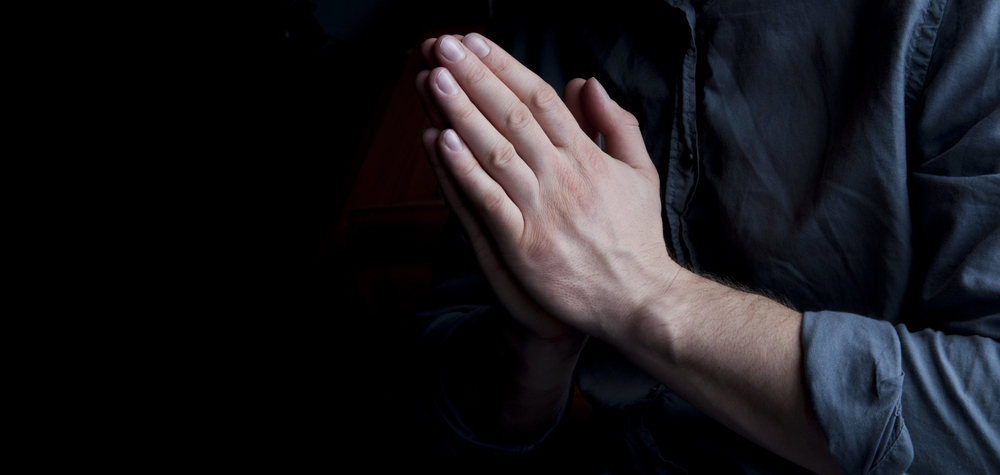 Hands pressed together in prayer against a dark background, person in a blue shirt,