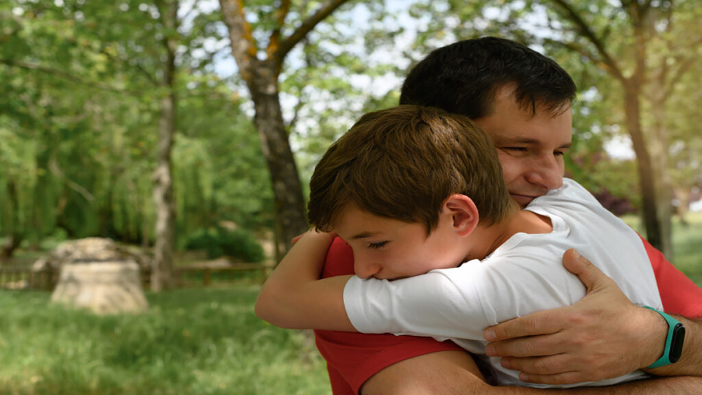 Father and son share a warm hug in a sunny park with green trees in the background.
