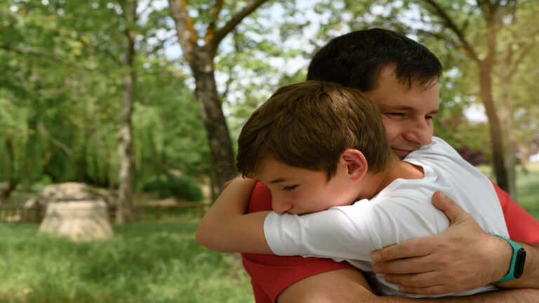 Father and son share a warm hug in a sunny park with green trees in the background.