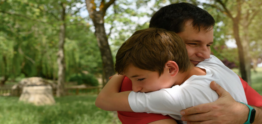 Father and son share a warm hug in a sunny park with green trees in the background.