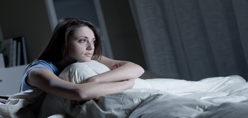 Woman resting on a bed with arms around a pillow, gazing pensively to the side in a dimly lit bedroom surface.