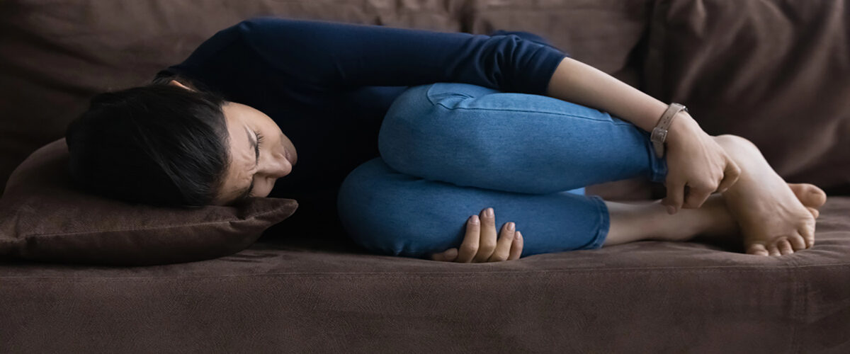 Person curled up on a brown sofa, knees to chest, head resting on a pillow, looking distressed.