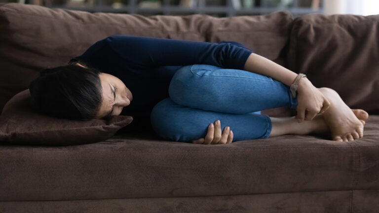 Person curled up on a brown sofa, knees to chest, head resting on a pillow, looking distressed.