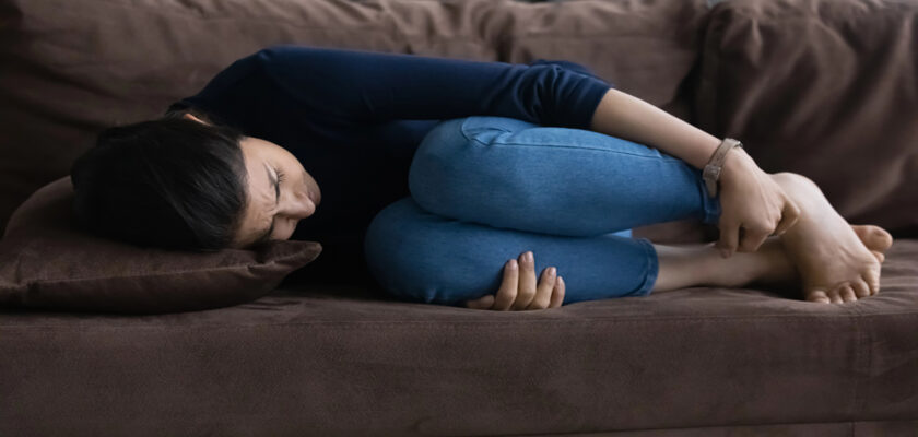 Person curled up on a brown sofa, knees to chest, head resting on a pillow, looking distressed.