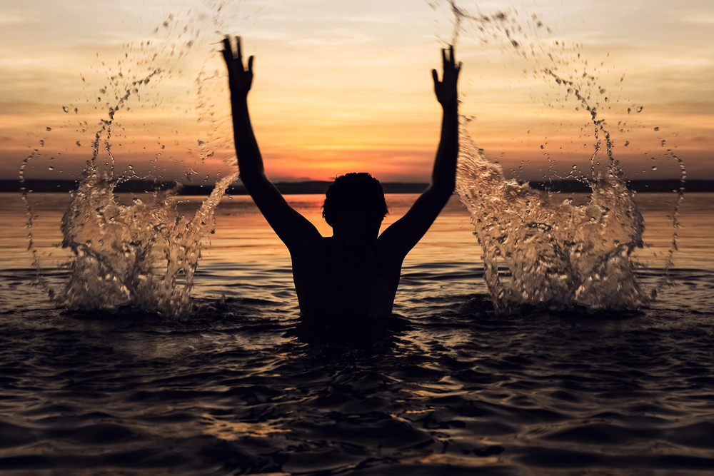 Depositphotos_506888666_S - Renuevo De Plenitud Silhouette of a person in water with arms raised, splashing as a sunset glows on the horizon.
