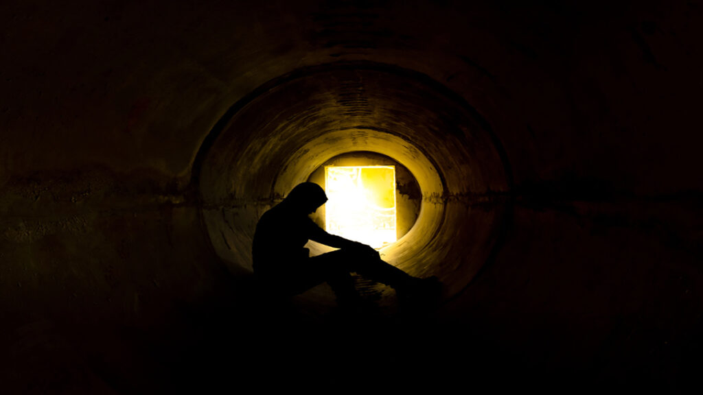 Silhouetted person sitting inside a dark circular tunnel, a bright exit glows at the far end.