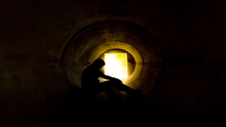 Silhouetted person sitting inside a dark circular tunnel, a bright exit glows at the far end.