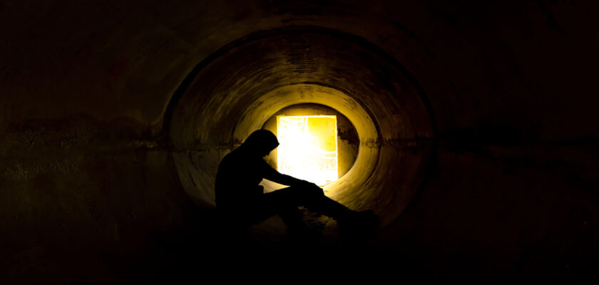 Silhouetted person sitting inside a dark circular tunnel, a bright exit glows at the far end.
