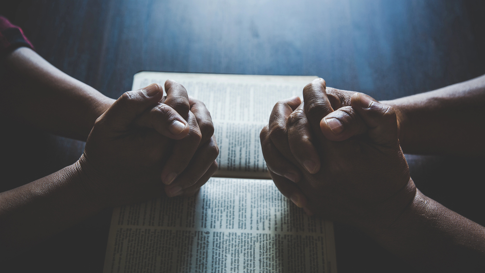 Depositphotos_530450898_S 1 - Renuevo De Plenitud Two pairs of hands clasped in prayer over an open book on a table, suggesting devotion or reflection.