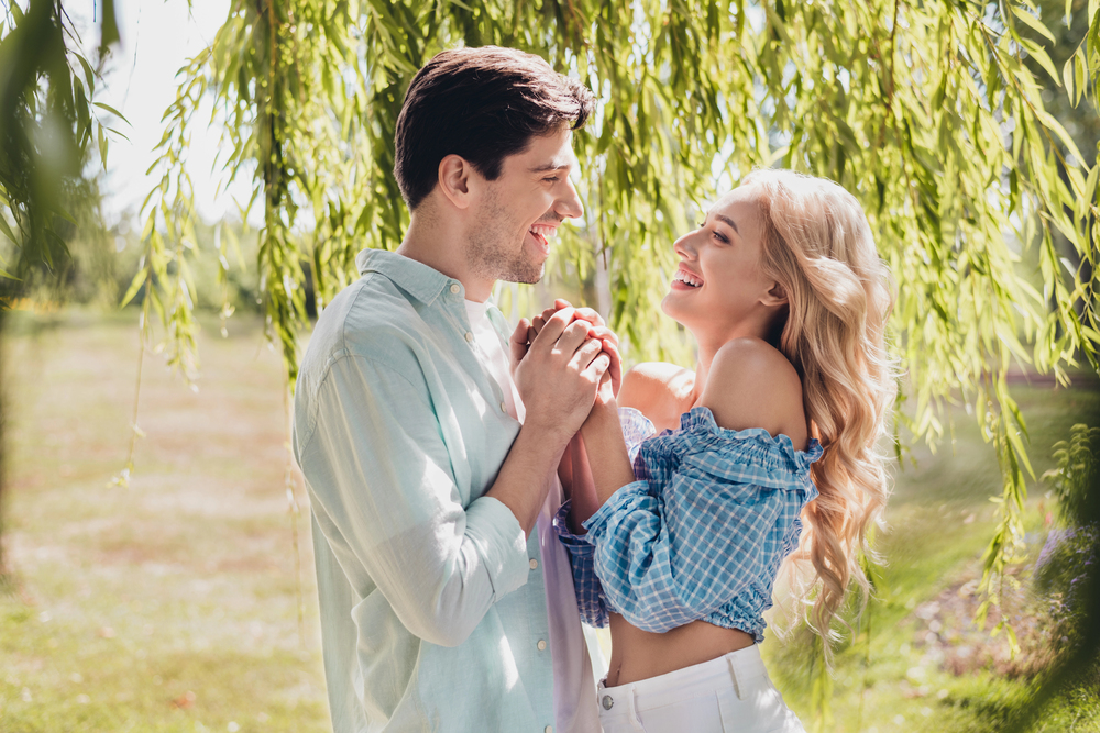 Young couple holding hands and laughing under hanging willow branches in a sunlit park.