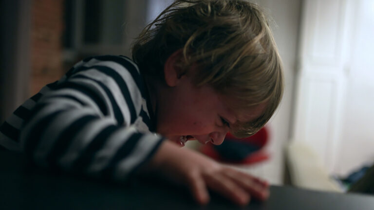 Young child in a striped shirt crying and leaning over a dark table indoors. ','A small child crying with a scrunched face, leaning toward a table. ','Little boy wearing a striped top crying at a table in a dimly lit room. ','Child at a table, distressed and crying, with a window behind. ','Young child expressing distress while gripping the edge of a table indoors.