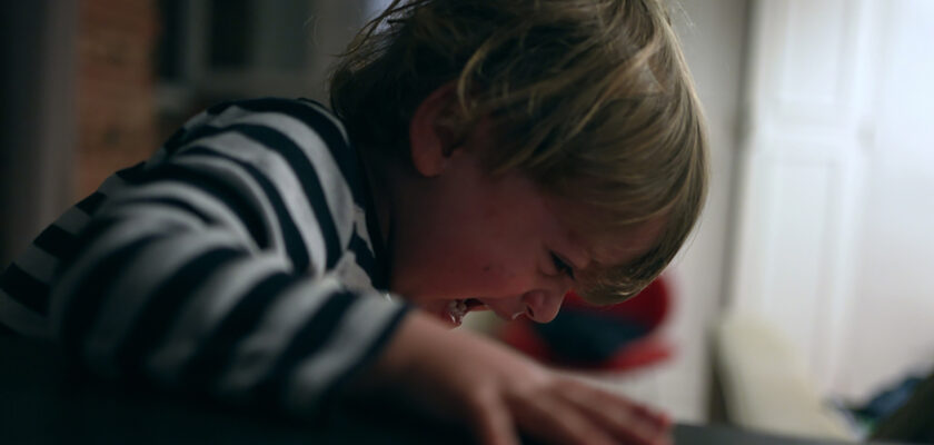 Young child in a striped shirt crying and leaning over a dark table indoors. ','A small child crying with a scrunched face, leaning toward a table. ','Little boy wearing a striped top crying at a table in a dimly lit room. ','Child at a table, distressed and crying, with a window behind. ','Young child expressing distress while gripping the edge of a table indoors.