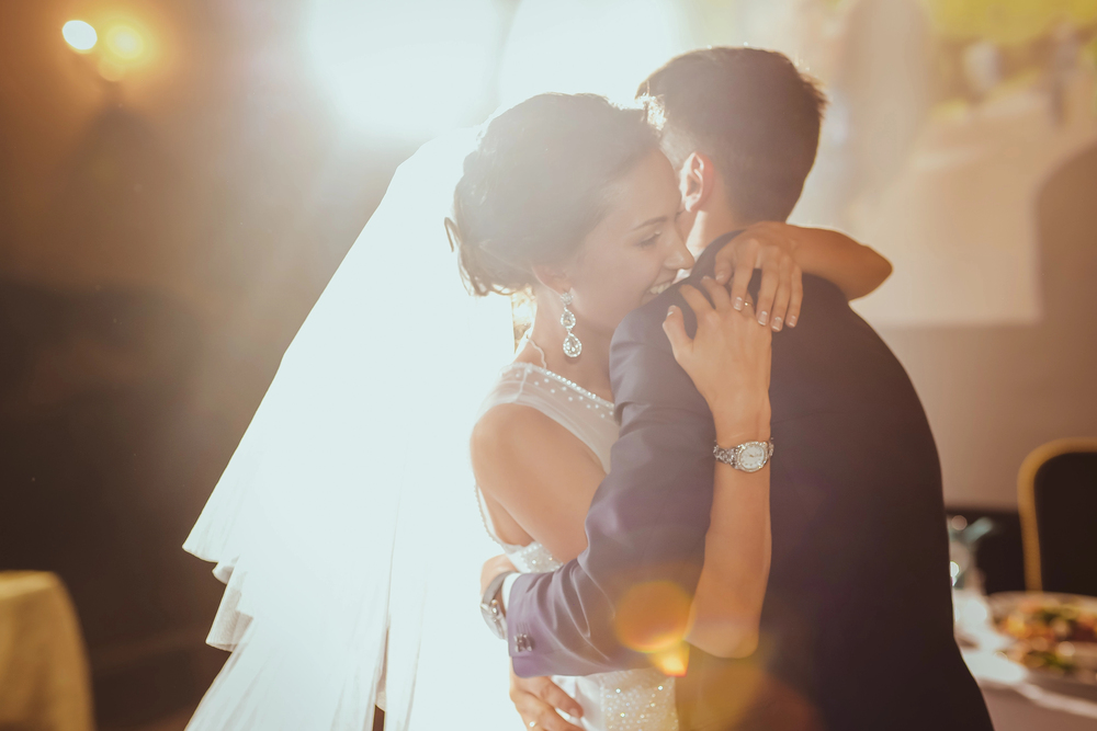 Bride and groom embrace and kiss during a wedding reception, backlit with warm lights in the background.