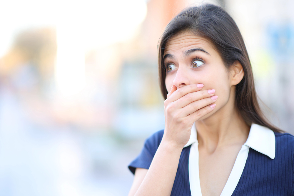A young woman outdoors covering her mouth in surprise or shock, eyes wide open.