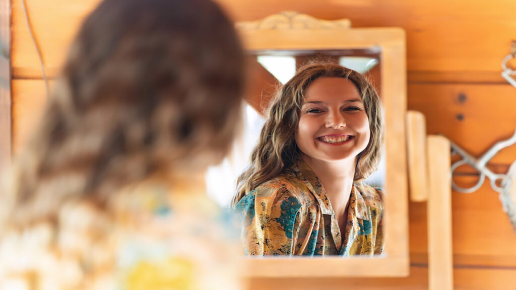 Smiling woman with wavy hair looks at her reflection in a wooden-framed mirror in a rustic room.