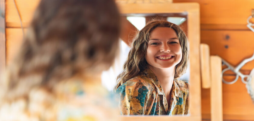 Smiling woman with wavy hair looks at her reflection in a wooden-framed mirror in a rustic room.