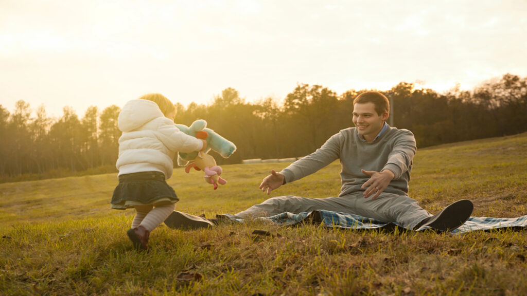 Father and young child playing in a sunlit park on a blanket, tossing plush toys to each other with smiles on their faces.