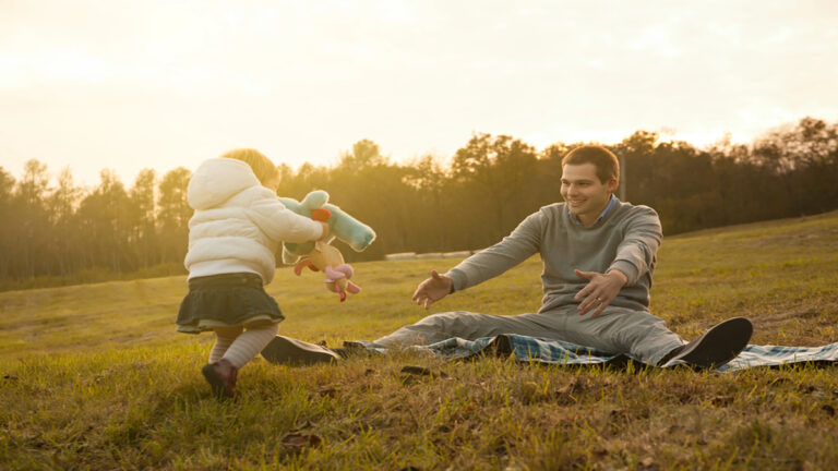 Father and young child playing in a sunlit park on a blanket, tossing plush toys to each other with smiles on their faces.