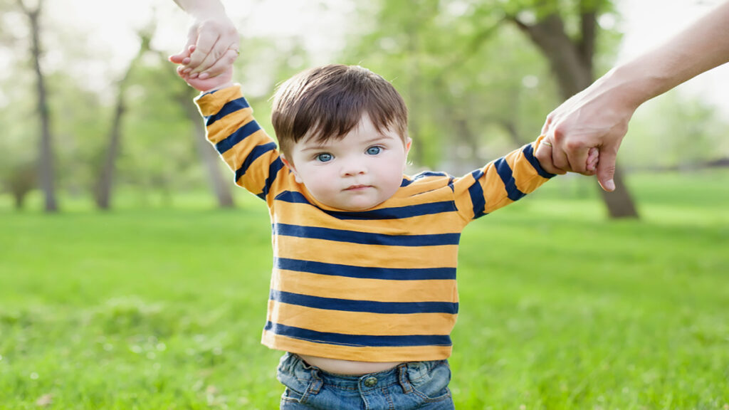 Toddler in a striped yellow and navy shirt is helped to walk in a sunny park, holding hands with two adults.