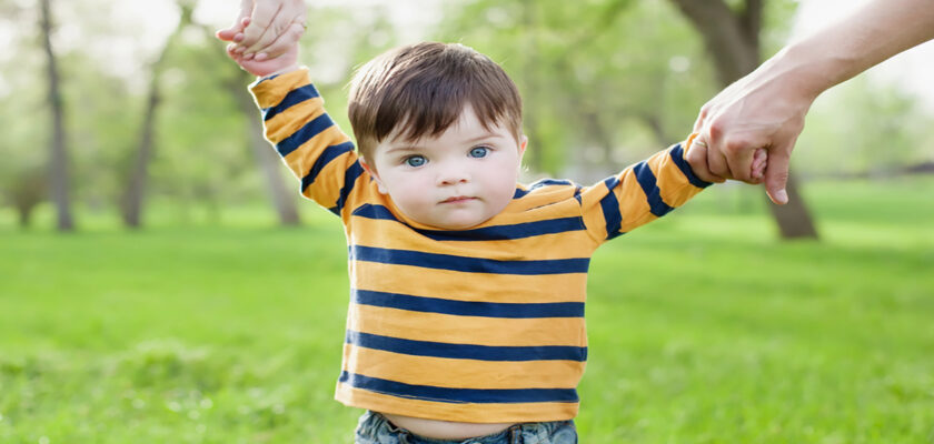 Toddler in a striped yellow and navy shirt is helped to walk in a sunny park, holding hands with two adults.