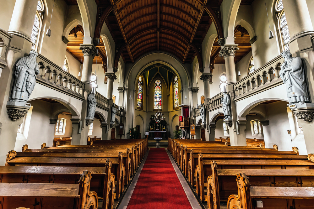 Interior of a church with wooden pews, statues along the walls, and a red carpeted aisle leading to the altar under stained-glass windows.