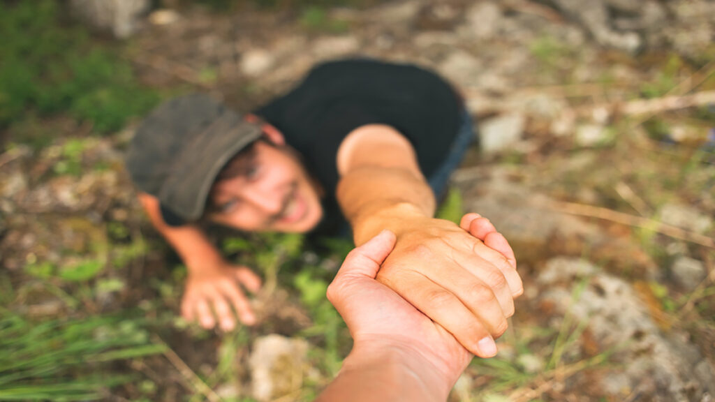 Person on the ground reaches up while another person grips their hand to pull them up in a rocky outdoor area.