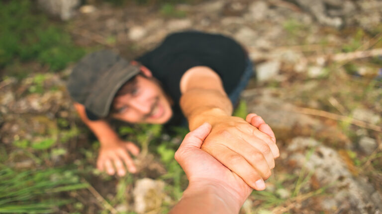 Person on the ground reaches up while another person grips their hand to pull them up in a rocky outdoor area.