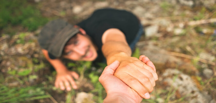 Person on the ground reaches up while another person grips their hand to pull them up in a rocky outdoor area.