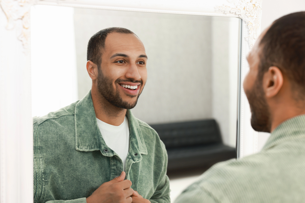Depositphotos_768695160_S - Renuevo De Plenitud Man with a beard smiles at his reflection in a large ornate-framed mirror, wearing a green corduroy jacket.