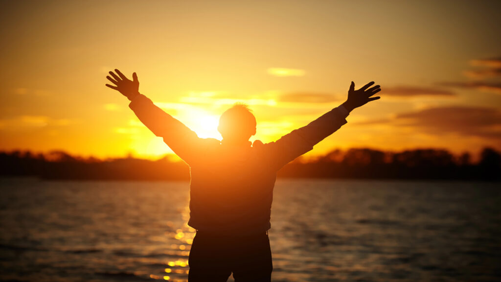 Person silhouetted with arms raised at the shore during a golden sunset over a calm lake/twater.