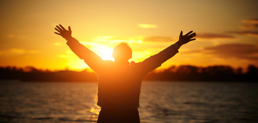 Person silhouetted with arms raised at the shore during a golden sunset over a calm lake/twater.