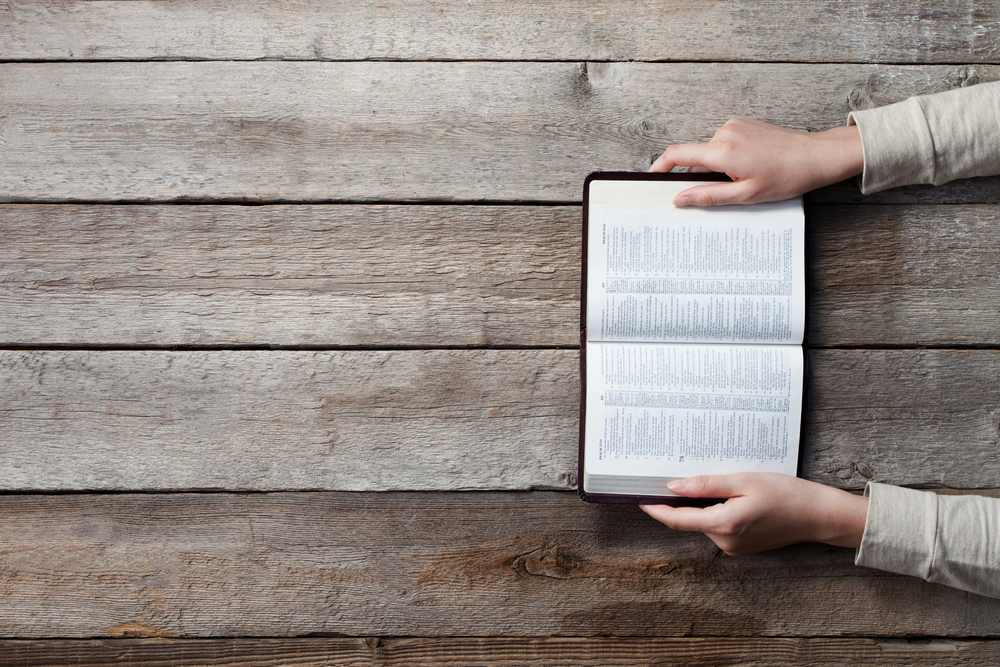 Top-down view of a person holding an open book with dense text on a weathered wooden table