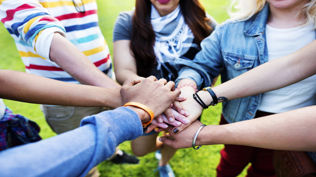 Group of friends placing hands together in a circle on a grassy field to symbolize teamwork and unity.